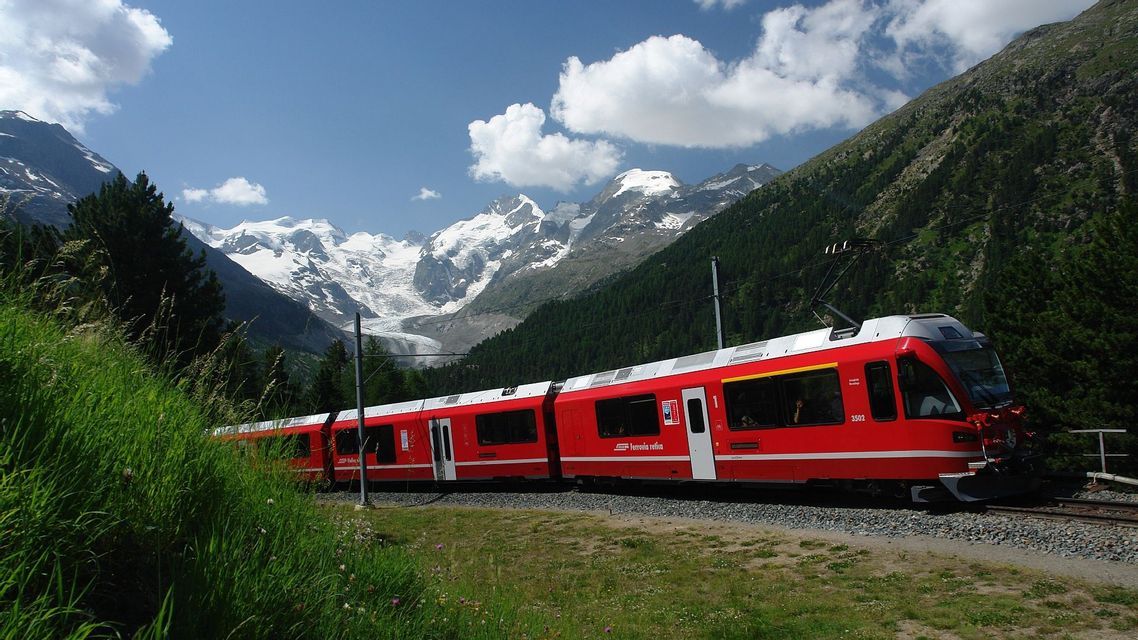 Un treno rosso viaggia su un binario attraverso una valle verde con uno sfondo di montagne innevate e un ghiacciaio.