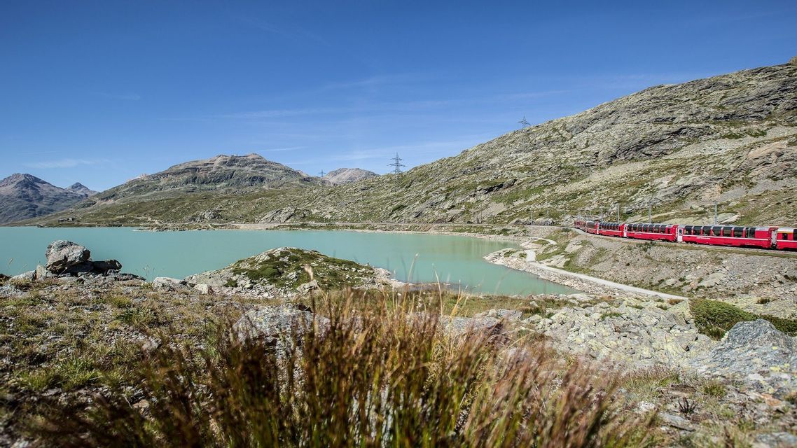 Un treno rosso viaggia su un binario lungo un lago alpino turchese e montagne rocciose sotto un cielo blu chiaro.
