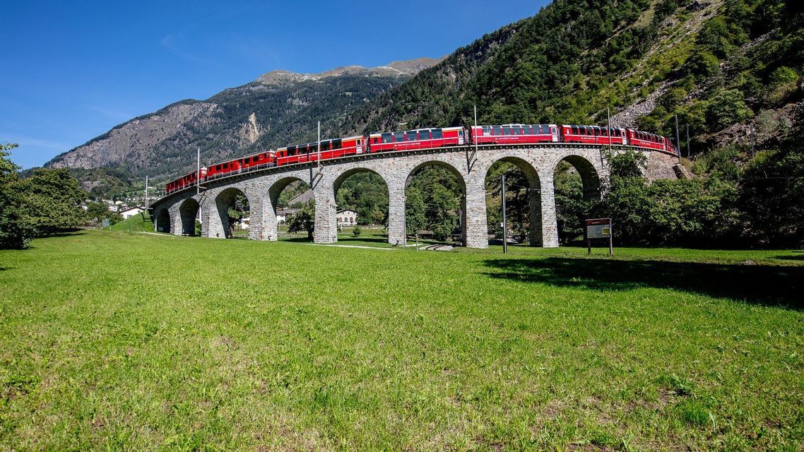 Un treno rosso attraversa un viadotto ad archi in pietra in una verde valle montana in una giornata di sole.