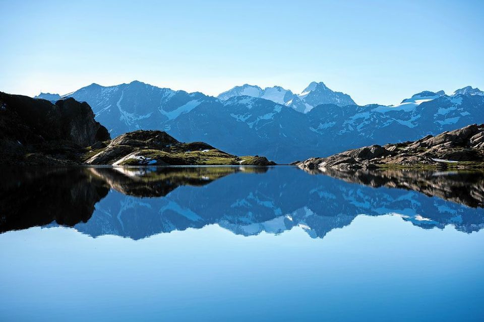 Una catena di montagne innevate che si riflettono perfettamente nelle acque calme di un lago alpino sotto un cielo azzurro limpido.