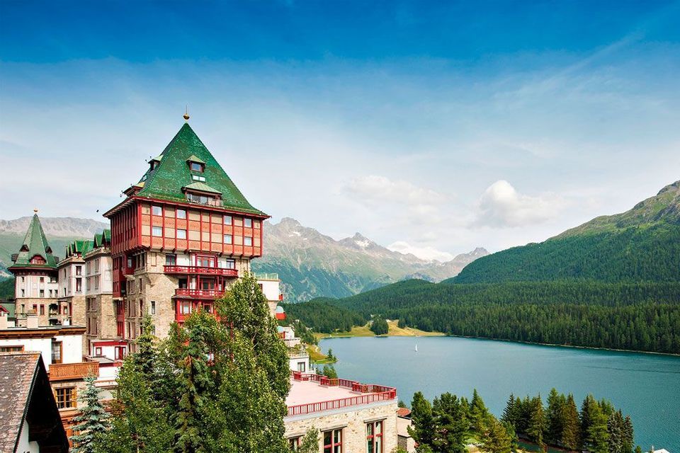 Un grande hotel con un prominente tetto verde si affaccia su un lago tranquillo, con montagne boscose sullo sfondo.