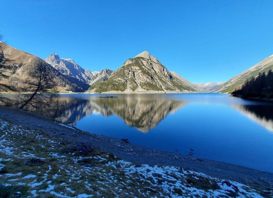 Montagne che si riflettono perfettamente in un lago alpino immobile con sponda innevata sotto un cielo azzurro limpido.
