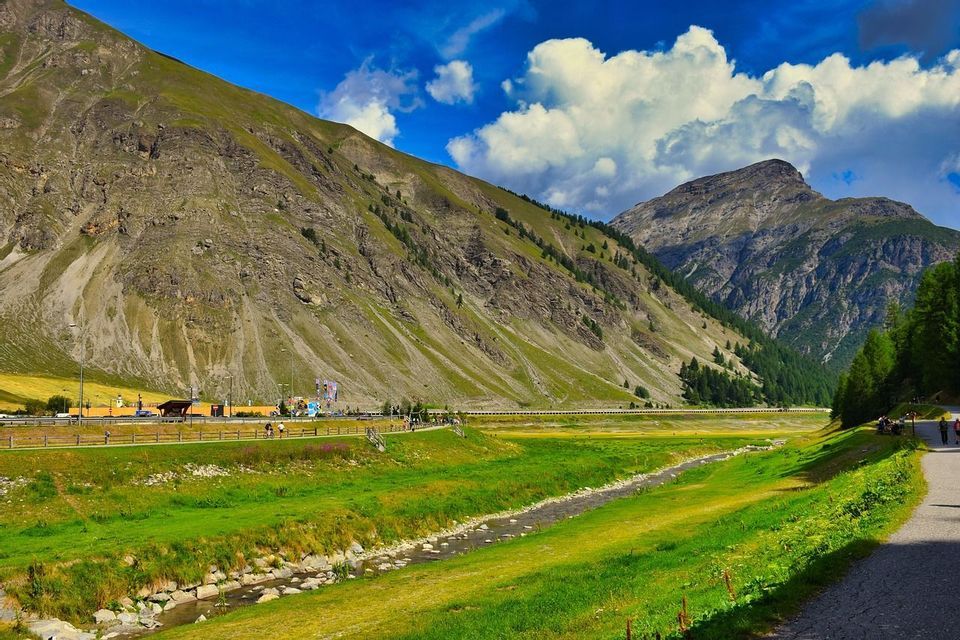 Un ruscello serpeggia attraverso una lussureggiante valle verde ai piedi di grandi montagne ripide sotto un cielo blu con nuvole bianche.