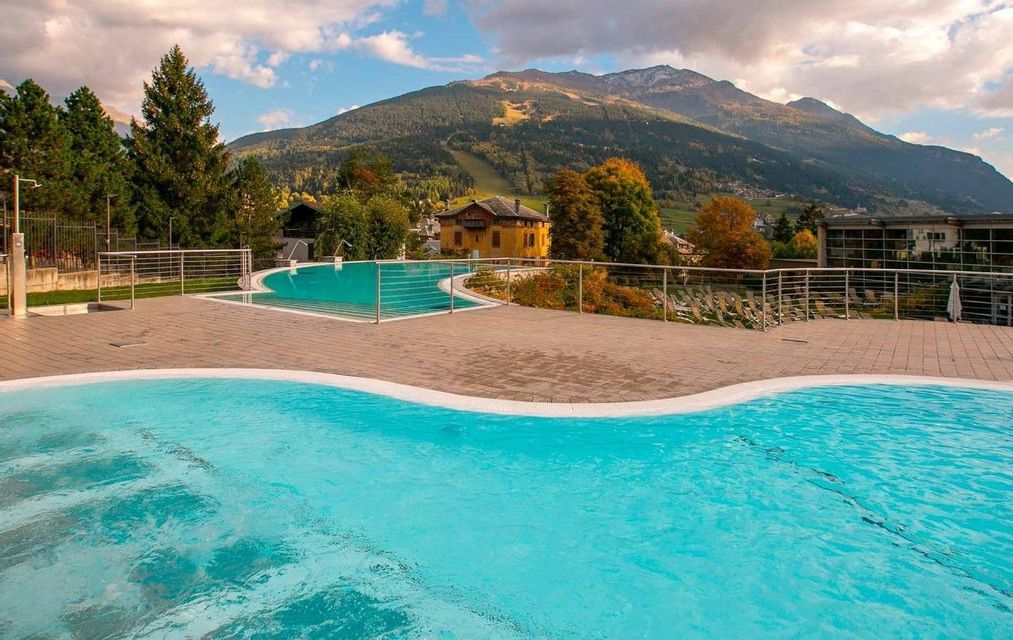 Una piscina all'aperto con acqua turchese su una terrazza pavimentata, con vista su una montagna boscosa sotto un cielo nuvoloso.