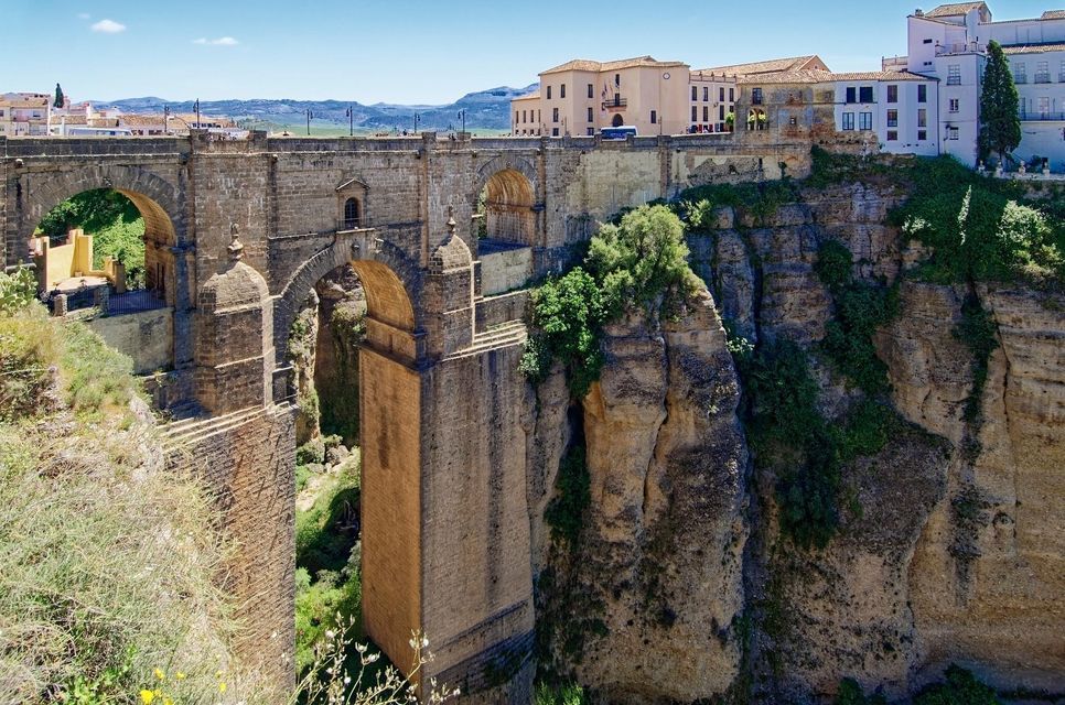 Un grande ponte in pietra con molteplici archi attraversa una gola profonda e rocciosa, con una città arroccata sulle scogliere sotto un cielo azzurro e limpido.