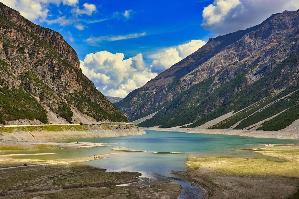 Un lago turchese si snoda attraverso una profonda valle tra ripide montagne rocciose sotto un cielo blu con nuvole bianche.