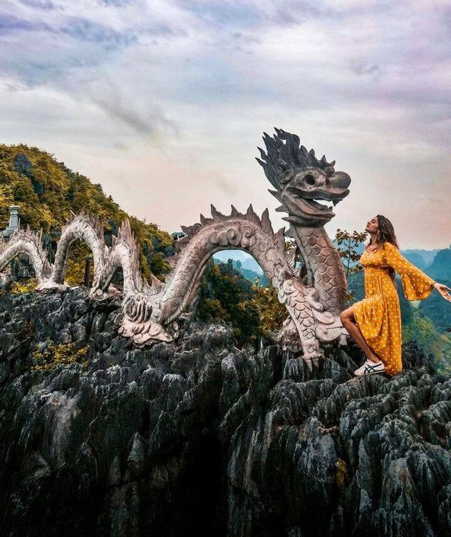 Una donna in abito giallo posa su una cima rocciosa accanto a una grande scultura di drago in pietra.