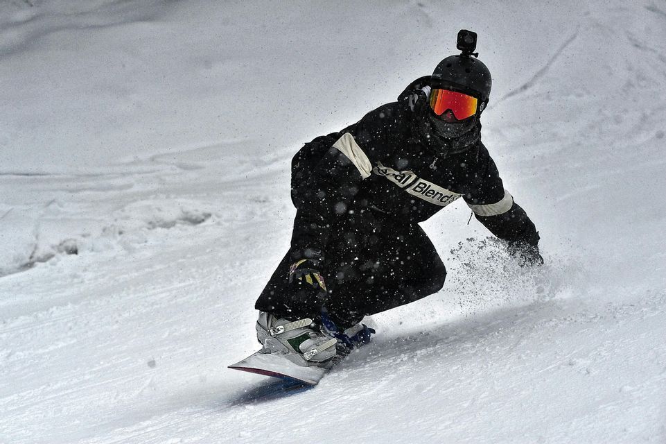 Uno snowboarder con casco e telecamera scende un pendio innevato, sollevando neve fresca mentre nevica.