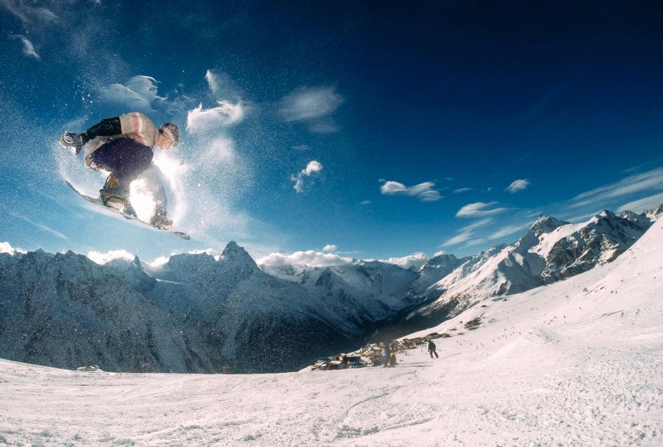 Uno snowboarder salta in aria sopra una catena montuosa innevata sotto un cielo azzurro chiaro.