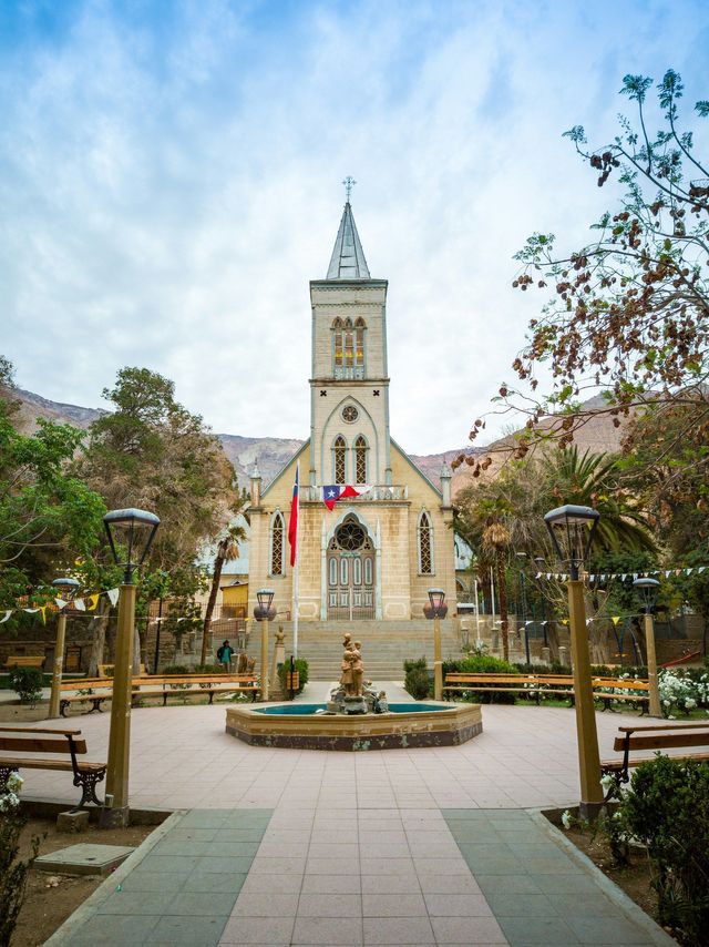 Una chiesa in pietra con un alto campanile e due bandiere si erge in una piazza con una fontana, circondata da alberi e montagne.