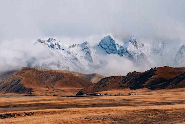 Snow-capped mountain peaks rise through clouds above a vast, golden steppe with a small settlement in the distance.