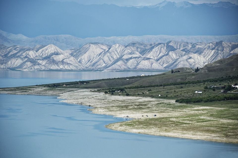 Un vaste lac bleu serpente le long d&#39;une rive de collines verdoyantes, avec des couches de chaînes de montagnes blanches et bleues en arrière-plan.