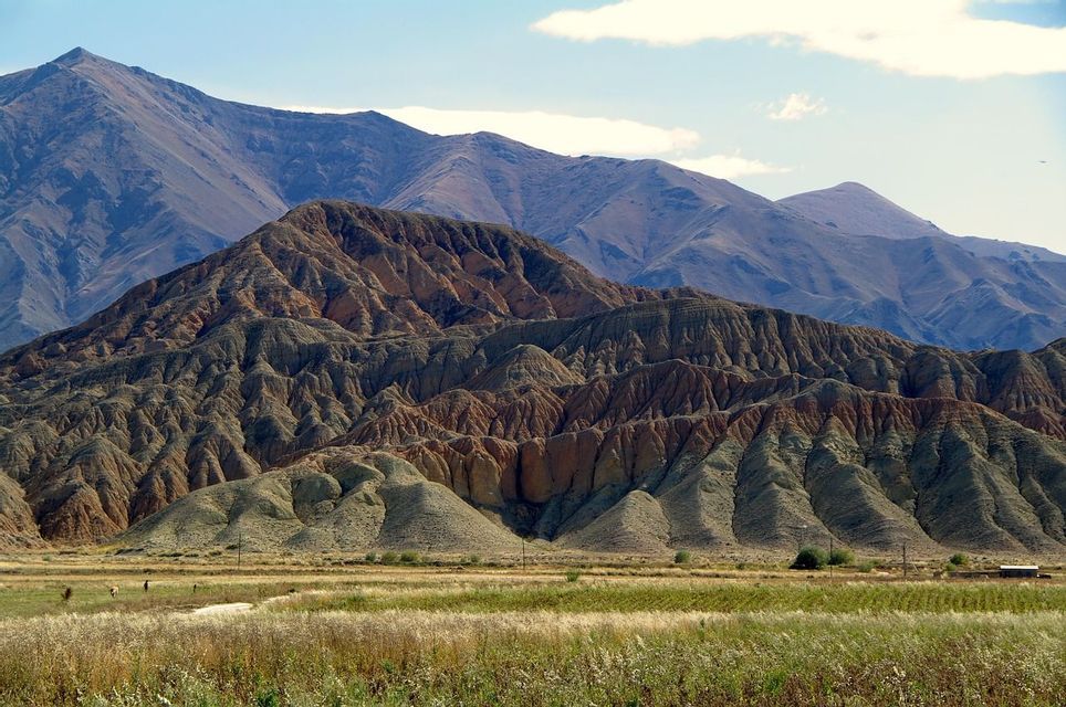 Des collines érodées avec des couches de roche rouge et verte se dressent devant une grande chaîne de montagnes, avec une plaine herbeuse au premier plan.
