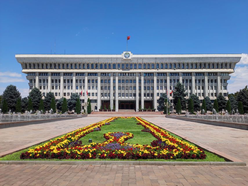 A large government building with a flag on top, viewed from across a plaza with fountains and colorful flowerbeds.