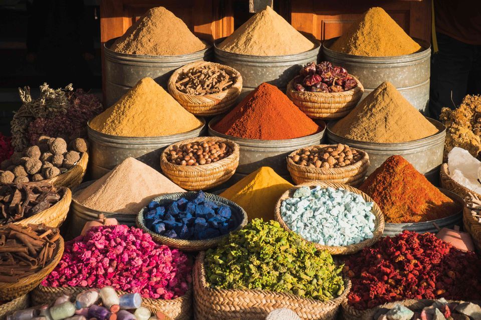 Spezie colorate in polvere e prodotti secchi disposti in mucchi e cesti intrecciati presso una bancarella del mercato all'aperto.