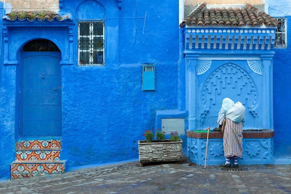 Una persona con abiti tradizionali a righe si china su una fontana pubblica blu e decorata in una strada acciottolata.