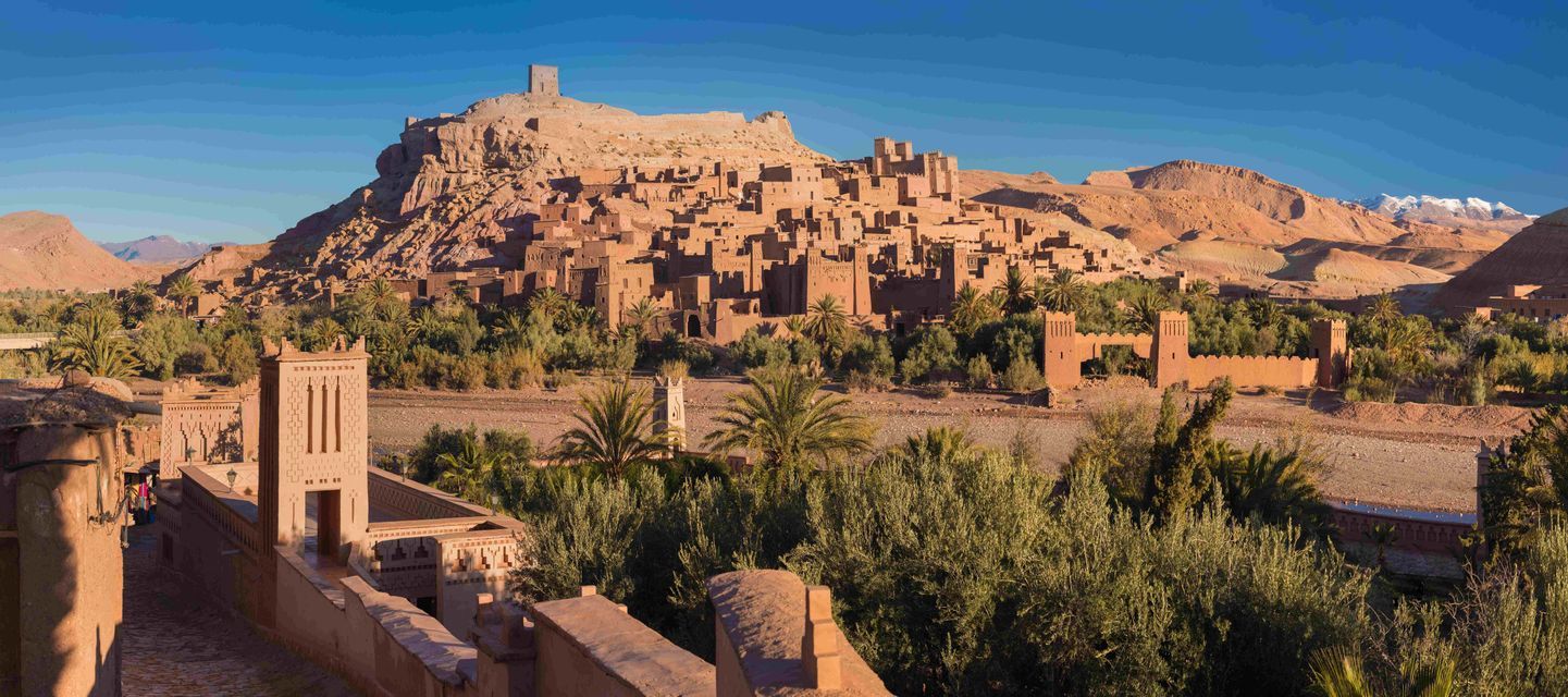 A panoramic view of a fortified mud-brick village on a hillside, overlooking a green oasis with distant, snow-capped mountains.
