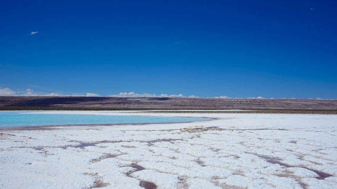 Una distesa di sale bianca con pozze d'acqua turchese si estende verso montagne lontane sotto un cielo azzurro limpido.