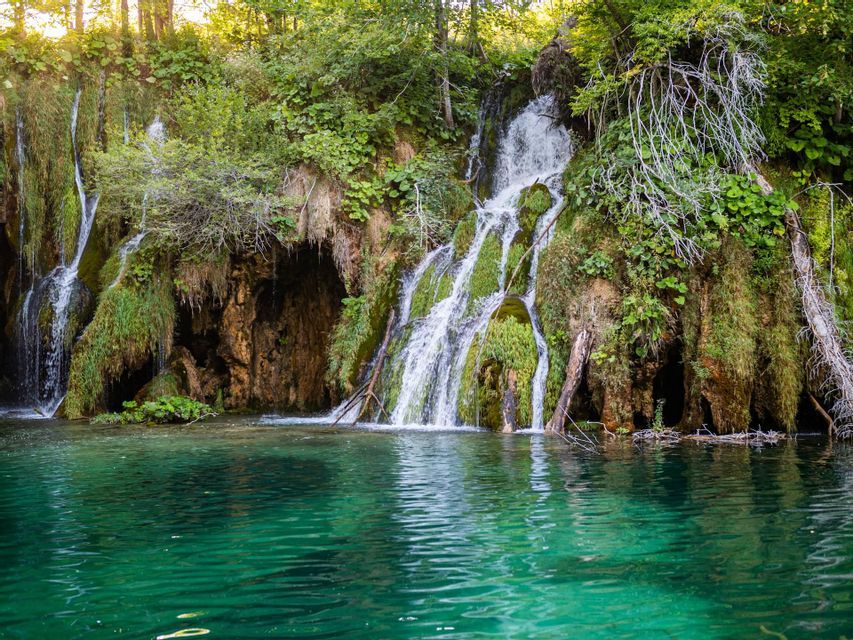 Wasserfälle stürzen einen moosbewachsenen Felsen hinab in einen türkisfarbenen See in einem sonnigen Wald.