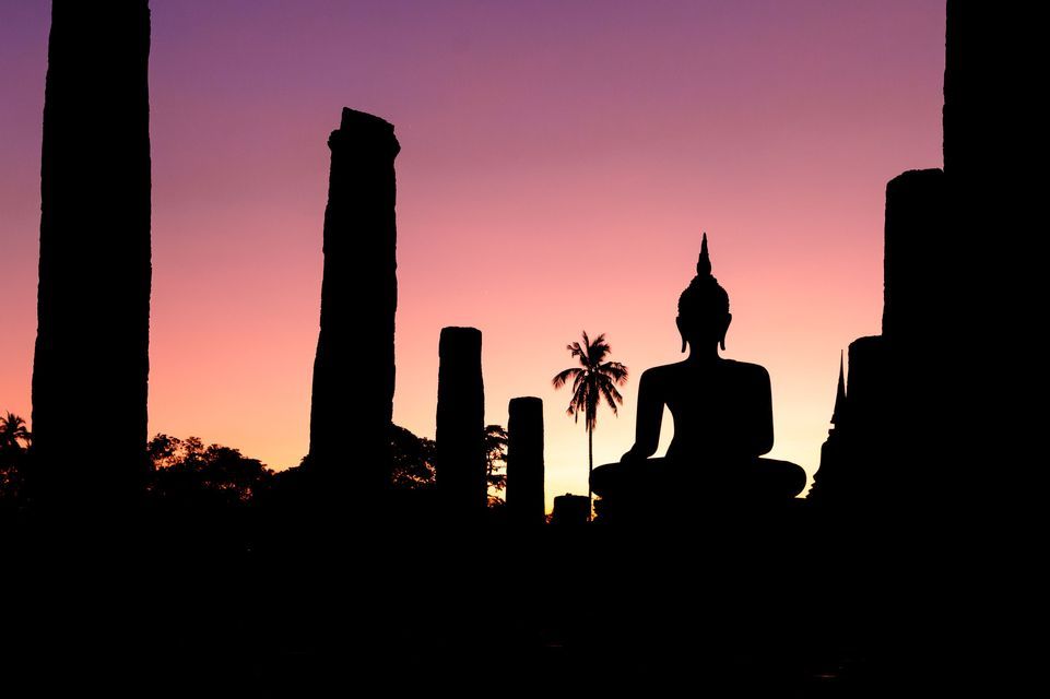 Una silhouette di una statua di Buddha seduto tra antiche rovine e una palma contro un cielo al tramonto viola e rosa.