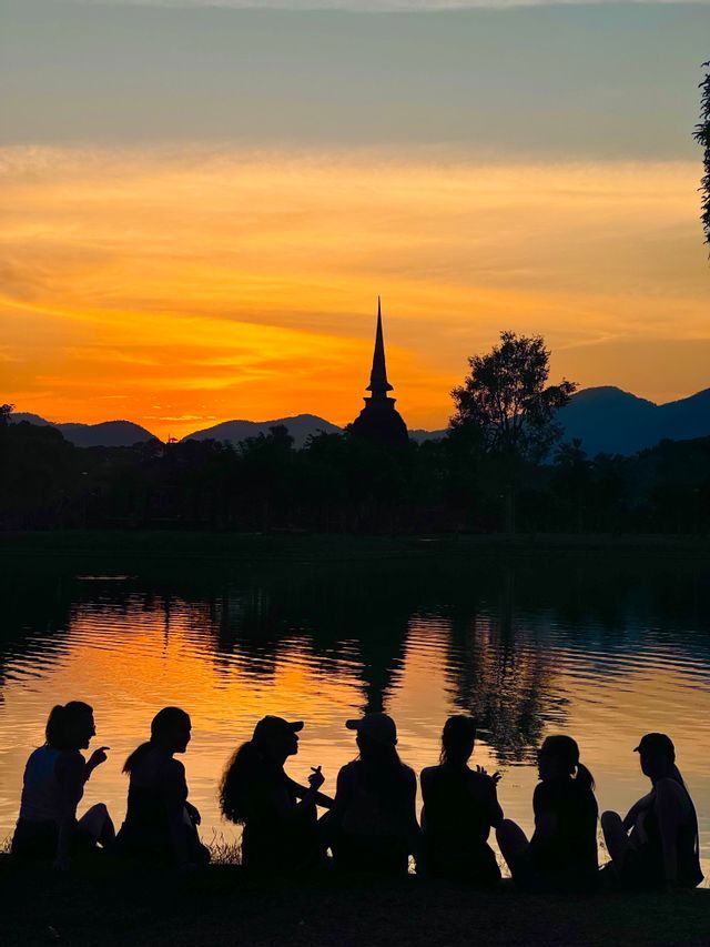 Eine WeRoad-Gruppe sitzt als Silhouette an einem See und beobachtet einen orangefarbenen Sonnenuntergang hinter einem fernen Tempel und Bergen.