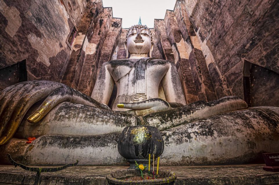 Eine kolossale, verwitterte Stein-Buddha-Statue sitzt meditierend zwischen hohen Steinmauern, mit einer kleinen Opferschale davor.