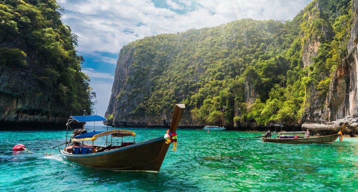 Zwei traditionelle Longtail-Boote treiben auf türkisfarbenem Wasser in einer Lagune zwischen steilen, bewaldeten Klippen.