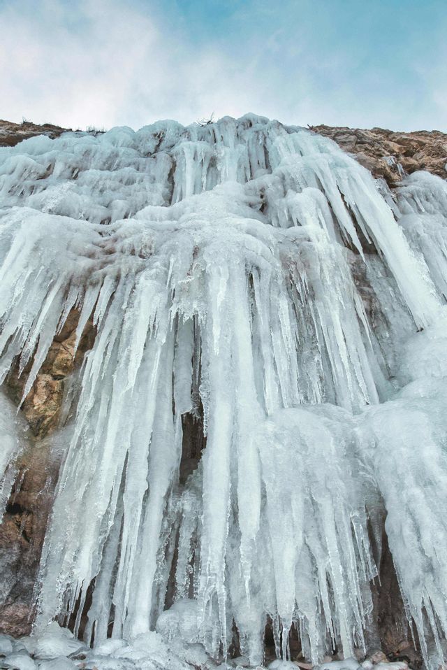 Eine Tiefenansicht eines gefrorenen Wasserfalls mit zahlreichen Eiszapfen, die an einer felsigen Klippenwand unter einem bewölkten Himmel hängen.