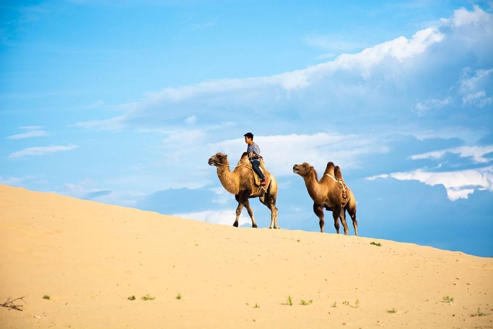 Eine Person reitet ein Kamel eine Sanddüne entlang, ein zweites Kamel begleitet sie, unter einem teilweise bewölkten blauen Himmel.
