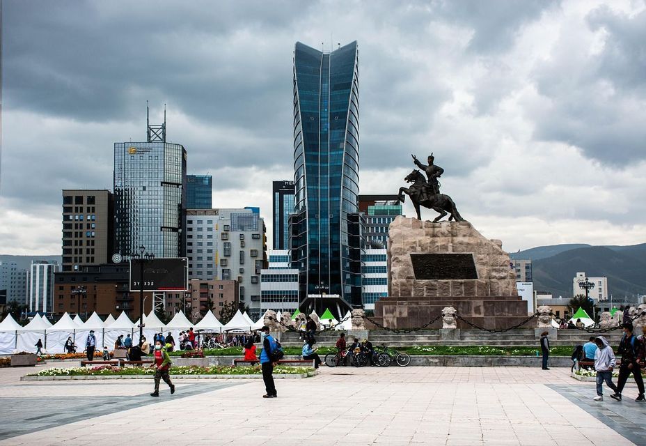 Eine bronzene Reiterstatue überblickt einen modernen Stadtplatz voller Fußgänger, mit gläsernen Wolkenkratzern und einem bewölkten Himmel im Hintergrund.