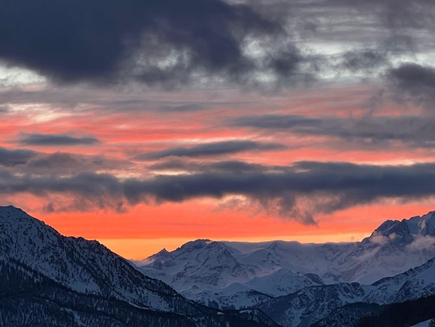 Un coucher de soleil coloré aux teintes orange et rose sous des nuages sombres illumine une chaîne de montagnes enneigée.