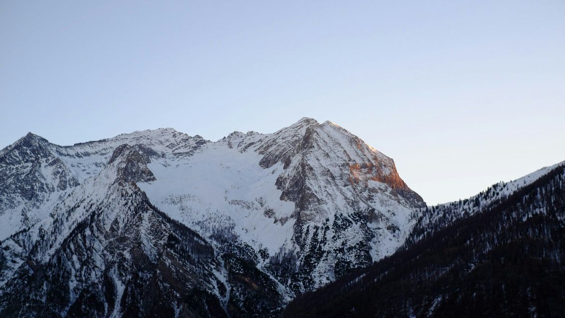 Un sommet de montagne enneigé capte la lumière dorée du soleil dans un ciel clair, s&#39;élevant au-dessus de pentes boisées sombres.