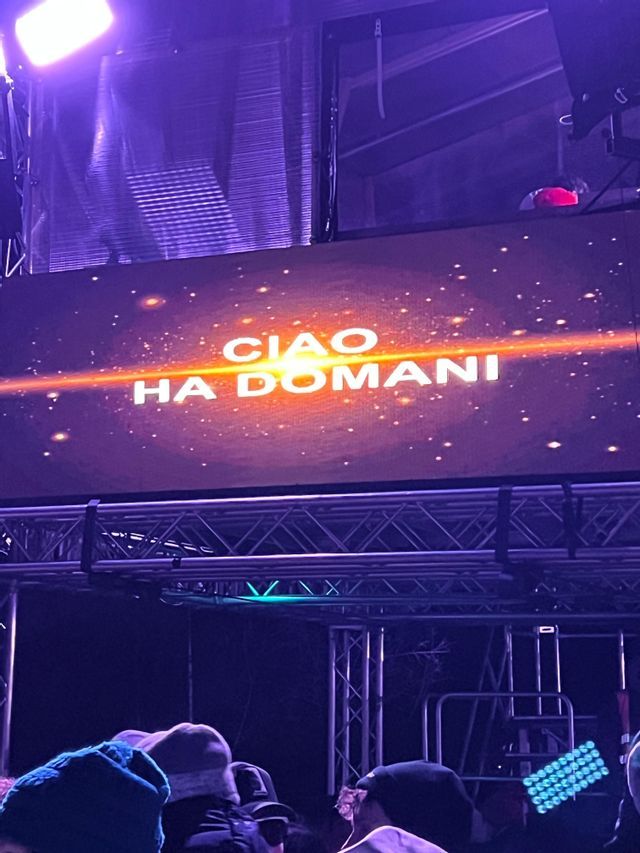 Un grand écran lors d&#39;un événement nocturne affiche le texte « CIAO HA DOMANI » sur un fond violet et cosmique, monté sur une structure de scène en métal.