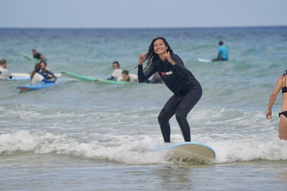 Une femme souriante lors d'un voyage de groupe WeRoad apprend à surfer, en équilibre sur sa planche dans l'océan avec d'autres.