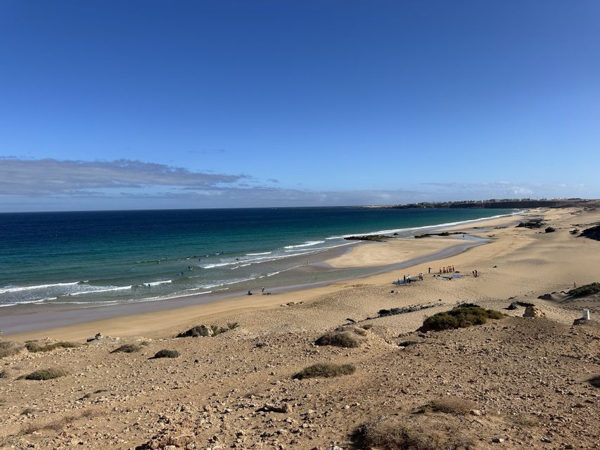 Un voyage de groupe WeRoad de surfeurs sur une plage de sable et dans l'océan turquoise, vu d'un point de vue élevé sous un ciel bleu.