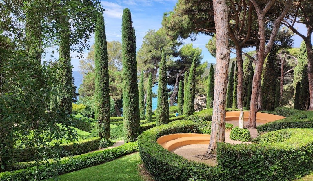 Un giardino rigoglioso con alti cipressi, siepi verdi scolpite e una panchina curva arancione che si affaccia sul mare.
