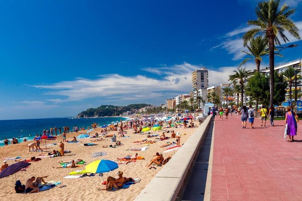 Molte persone prendono il sole su una spiaggia sabbiosa affollata accanto a un lungomare cittadino con palme sotto un cielo azzurro limpido.