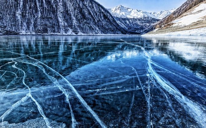 La glace bleue claire d&#39;un lac gelé est fissurée, reflétant une forêt et des montagnes enneigées.