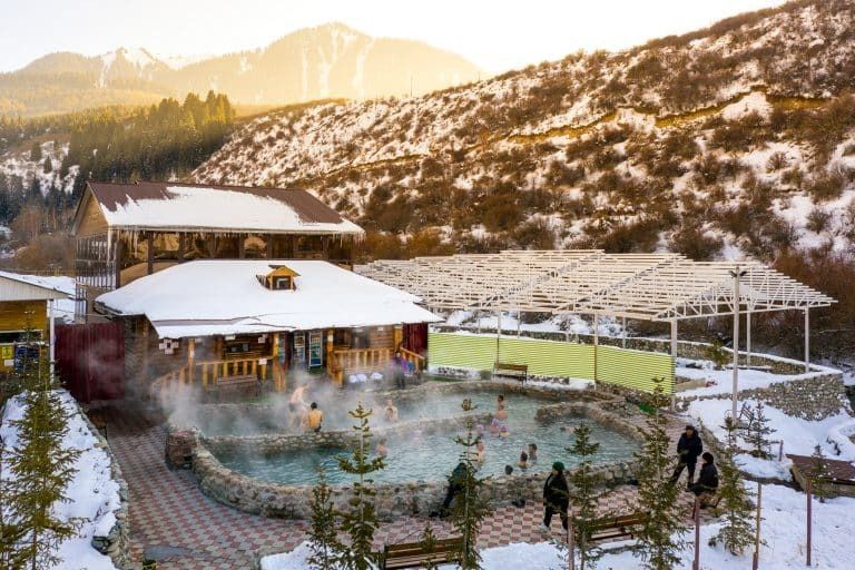 Un groupe WeRoad se détend dans une source chaude extérieure fumante, entouré de cabanes et de montagnes enneigées.