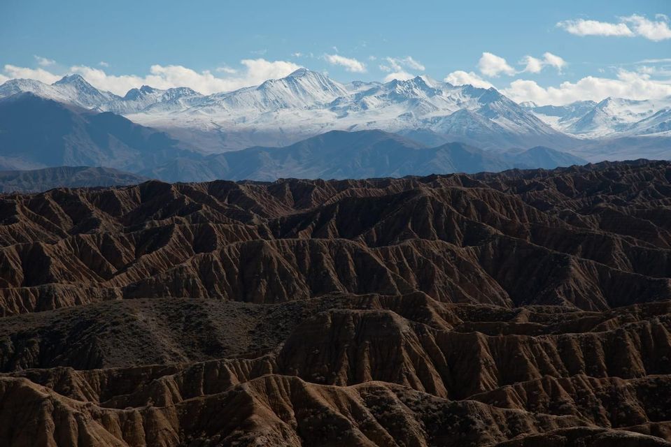 A landscape of brown, eroded hills in the foreground with a range of snow-capped mountains in the distance under a blue sky.