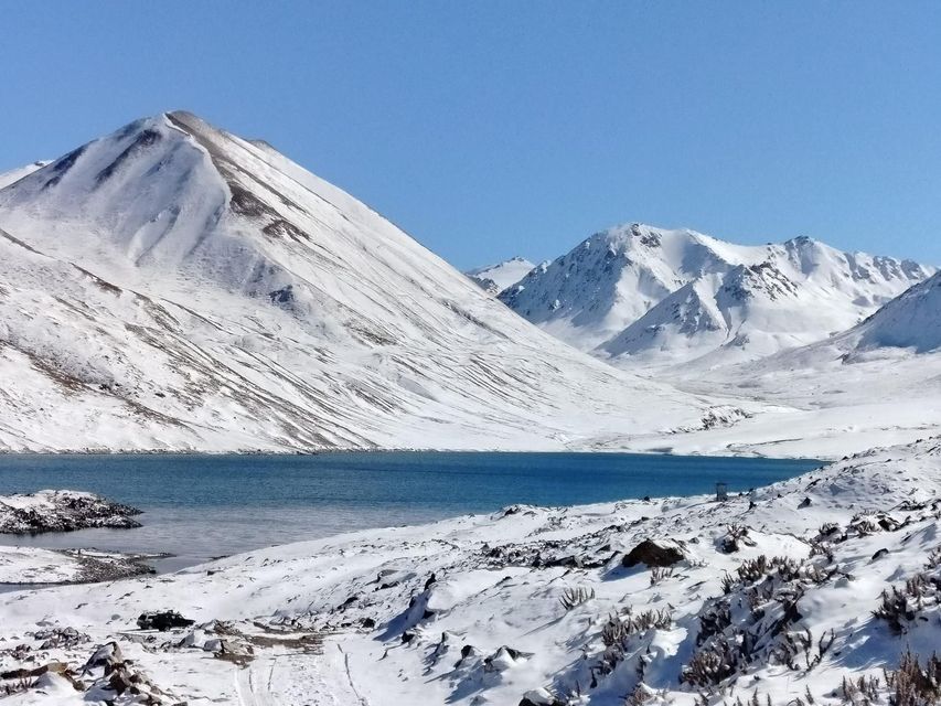 A blue alpine lake sits at the base of large, snow-covered mountains under a clear blue sky.