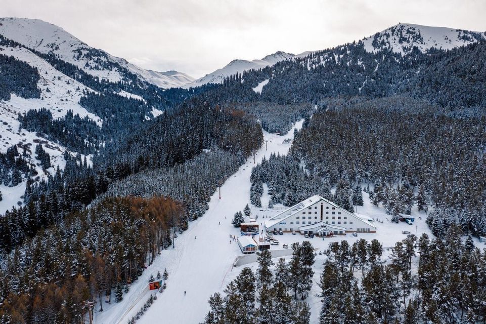 Vue aérienne d&#39;une station de ski avec un grand hôtel au pied d&#39;une piste, entouré de montagnes enneigées et de forêts de pins.