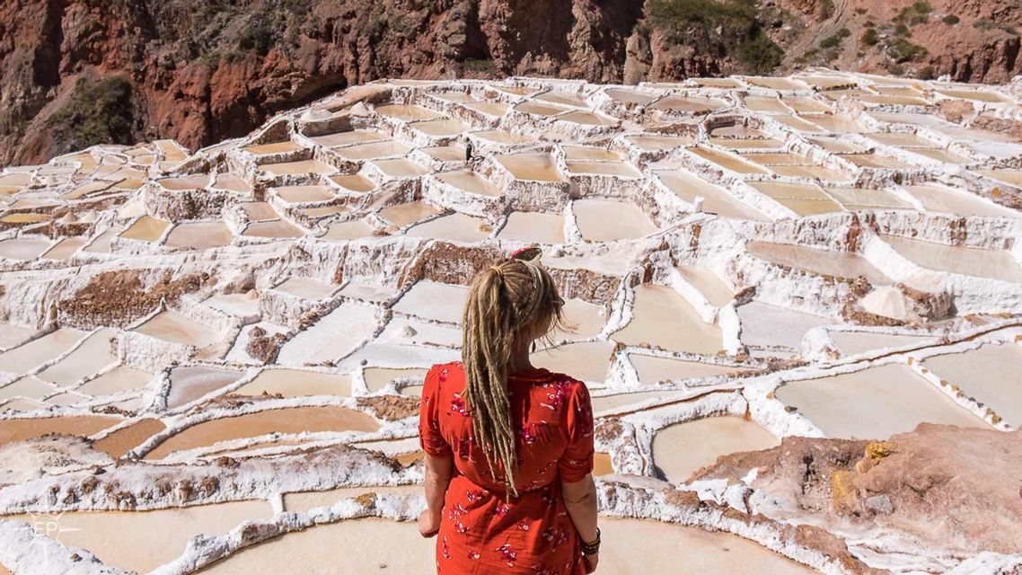 A woman with blonde dreadlocks in a red top, seen from behind, looking out over terraced salt evaporation ponds.