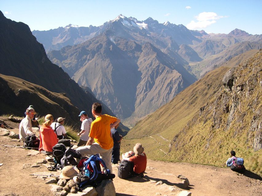 A WeRoad group trip taking a break on a mountain trail, overlooking a deep valley with distant snow-capped peaks.