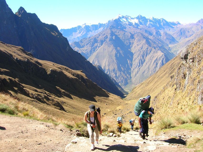 A WeRoad group trip hikes up a steep mountain trail, with snow-capped peaks visible in the background under a clear sky.