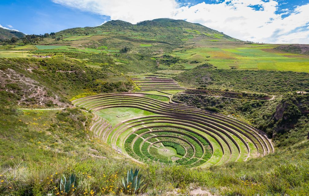 Concentric circular agricultural terraces are carved into a green valley surrounded by rolling hills under a blue sky.