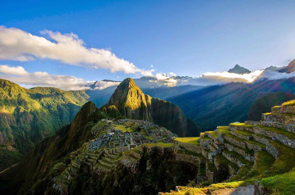 Sunlight hits the peak of a high mountain overlooking ancient stone ruins built into the lush, green mountainside.