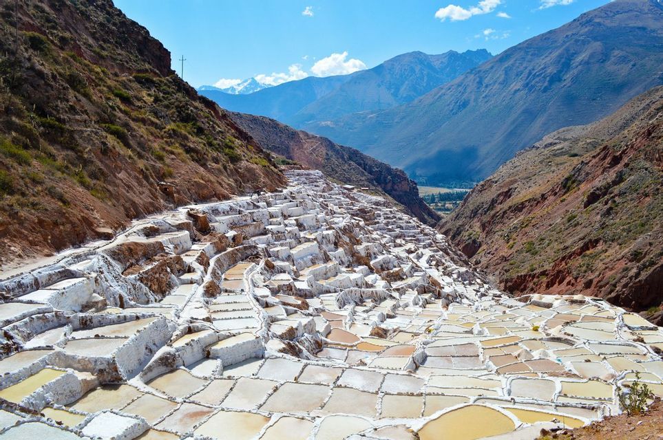 Terraced salt evaporation ponds cascade down a steep mountainside in a valley, with distant mountains under a blue sky.