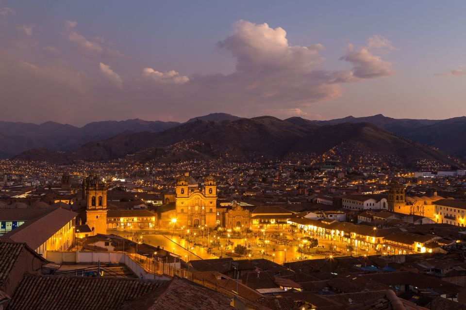 An illuminated city with a central plaza and cathedral at dusk, nestled in a valley surrounded by mountains.