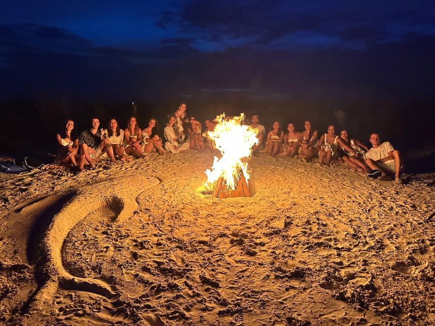 A WeRoad group trip gathered around a bonfire on a beach at night, with a large dolphin drawn in the sand in the foreground.